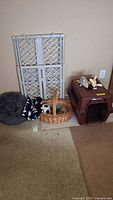 Photo showing folding pet gate, pillow bed, wicker basket with toys, and brown pet carrier crate arranged on carpeted floor against a beige wall.