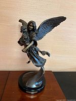 Front view of the bronze guardian angel sculpture showing the angel holding a child, with detailed wings and flowing robes on a black base.