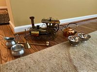 Photo showing the music box shaped like a train engine, a dented copper kettle, two pewter bowls (cream and sugar), a small brass bell, and a candle snuffer with wooden handle placed on the floor.