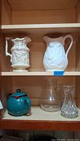 Two shelves in wooden cabinet featuring six items: two pitchers on top shelf, tea pot, glass pitcher, and glass vase on bottom shelf
