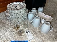 Stack of 10 large and 10 small plates with gold swirl design, 5 white Bistro Blanc mugs upside down, glass salt and pepper shakers, white sugar dish with text, and white creamer on carpeted floor.
