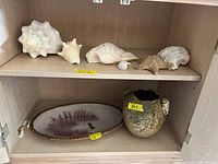 Shelf showing five assorted natural sea shells of various sizes and shapes on top shelf, signed 16-inch platter and textured pottery vase with curled handles on bottom shelf