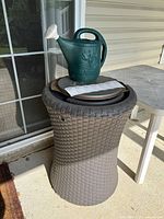 Photo of plastic rain barrel with woven wicker texture, starburst-style top, and green watering can along with plastic trays stacked on top, situated outdoors next to sliding glass door.