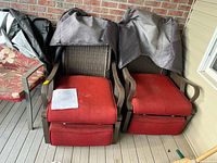 Two brown all-weather wicker patio reclining chairs with red removable cushions, with protective covers draped over the backrests, placed on covered deck flooring near brick wall