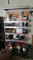 Full frontal image of metal and wood garage shelf unit showing five shelves with various items including helmets, lights, and small containers. Shelf metal frame with wood particle board shelves.