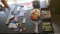 Table display showing religious icon plaque, stained glass angel ornament, various crosses, magnets, thermometer, and a box containing ornaments.