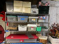 Photo showing full storage shelving unit with four shelves containing numerous plastic bins, drawers, boxes, tins, and foldable files, plus floral artificial plants nearby.