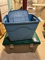 Two large plastic storage bins stacked in a basement. Blue bin on top with lid inside it, green-beige bin underneath with latches visible.