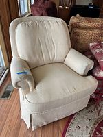 Front-left view of beige Jessica Charles swivel rocker chair showing cushioned back, rolled arm, seat cushion, skirted base, and discoloration on the arm.