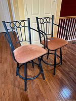 Two black metal swivel stools with rust orange cushioned seats, one with armrests and one without, on a wooden floor near a door.