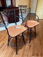 Front angle of two metal swivel stools with cushioned orange seats, black frames with armrests and decorative backs.