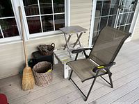 Outdoor deck scene showing folding chair, white and gray side tables, broom, baskets, watering can, and trash can in grouping near windowed wall.