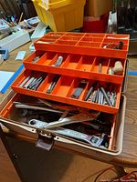 Photo showing the open orange tackle box with multiple trays containing hand tools including wrenches, pliers, screwdrivers and drill bits.