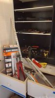 Photo showing various hand tools stored in a cabinet and drawers including saws, clamps, hammers, measuring instruments, and a boxed Black & Decker laser tool.