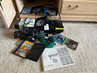 Overview photo showing PlayStation 2 slim console, controllers, game discs, and user manual on carpeted floor near wooden furniture.