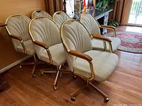 Six beige upholstered office chairs with wood armrests and gold metal frames, arranged in rows on a wood floor near windows and furniture.