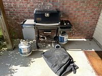 Full front view of Weber Genesis Silver propane grill with two propane tanks on either side and black grill cover on the ground in front of it.