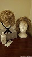 Two short wigs on stands and mannequin head with a white hair care bottle on a wooden surface.
