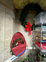 Two Christmas wreaths hanging in basement with a shelving unit and other storage items nearby. One wreath is large with red bow and string lights (untested) visible, and a smaller wreath in a red storage bag below.