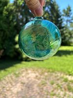 Full view of green iridescent handblown ornament held by loop outdoors