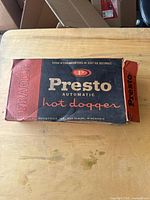 Photo of vintage 1940s Presto Automatic Hot Dogger in original cardboard box on wooden surface.