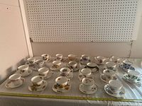 Overview of 24 assorted porcelain cups and saucers laid out on a white cloth surface showing variety of floral patterns and styles.