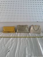 Photo showing all three vintage clutch handbags lined up on white cloth with measuring tape, from left to right: yellow clutch, metallic gold clutch, cream white clutch.
