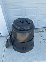 Full front and side view of black plastic composter placed on concrete in front of garage door, showing cylindrical vented body and lid with handle.
