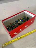 Top-side view of red box containing assorted glass marbles of varying colors and sizes, with a measuring tape beside it showing the box length around 12 inches.