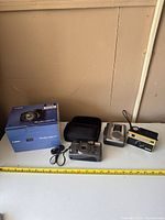 Four items on table including Canon Sure Shot 85 Zoom in carrying case, Kodak Instant Load 900 camera, small point and shoot camera, and cassette recorder with tape deck.