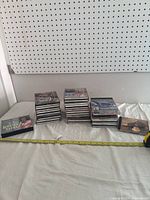 Multiple stacks of classic country music CDs on a white surface against a pegboard background. A yellow measuring tape is placed in front.