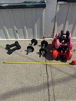 Overview of all dumbbells included in the lot arranged on concrete ground showing two black neoprene dumbbells, two adjustable dumbbells with metal plates, and a stack of six fixed-weight pink and red neoprene dumbbells.