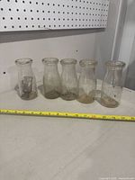 Five old half pint glass milk bottles arranged side by side with measuring tape showing approximate dimensions