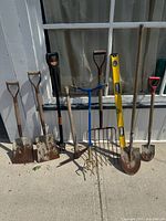 Full view of all the garden tools in lot including shovels, pick axe, digging fork, spirit level, and hoe.