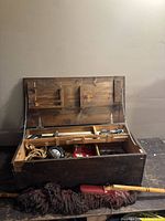 Wooden tool chest open showing internal compartments with tools including mop, rope, saw, tape measure, and other tools inside.