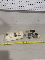 Original labeled box with mineral slots visible and some loose mineral rocks next to it on table. Measuring tape used for scale.