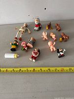 Overview of all 14 miniature dolls and figures arranged on a flat surface with measuring tape for scale. Items include baby dolls, cartoon characters, Santa Claus ornament, and a plastic baby bottle.