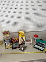 Wide shot showing all games and toys in the lot arranged on a floor against a pegboard wall. Visible items include Mastermind, Spill & Spell, Currency Cash toy, King Corner cards, WWE electronic game, and domino set.
