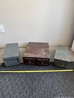 Three old index boxes, two metal (gray and green) and one wood with two drawers, side by side on carpeted floor against wall.