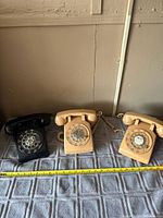 Three rotary dial phones: two beige and one black on gray cloth with measuring tape showing approx 8 inches