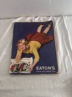 Front cover photo of the 1944 Eaton’s Spring and Summer Catalogue showing a woman lying on her stomach wearing a yellow sweater and red plaid skirt, holding the catalogue open.