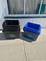 Ten plastic recycling bins in black and blue, stacked outside on pavement against a wall
