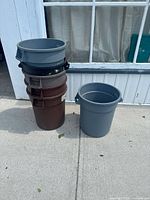 Six plastic garbage cans stacked and placed on sidewalk, mixture of brown and gray colors, showing surface wear.