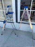 Two aluminum step ladders standing open on a sidewalk outside a building with windows, showing overall structure and appearance