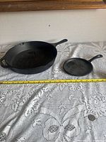 Two cast iron pans placed side by side on a lace tablecloth with a measuring tape showing size comparison and a coin for scale.