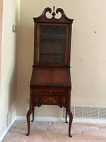 Front view of antique wooden secretary desk showing glass-front upper cabinet with shelves, intricately carved top finial, drop-front desk in closed position, and three drawers below with metal ring pulls.