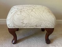 Front view of the upholstered ottoman showing beige damask fabric with a large leaf floral pattern and brown wooden cabriole legs.
