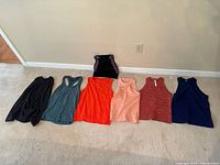 Six women's sleeveless tops laid out: black, teal, orange, peach, brick red, navy blue, plus one black and purple sports bra and one black fleece sweatshirt.
