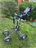 Three angled photos of the black and gray Bag Boy Quad golf push cart standing on grass with a green garden background. Shows details of the four wheels, handle with foam grip, storage pouch, and brand labeling.
