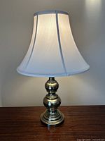 Full view of the lamp assembled and lit with ivory shade and brass base on wooden surface against plain background.
